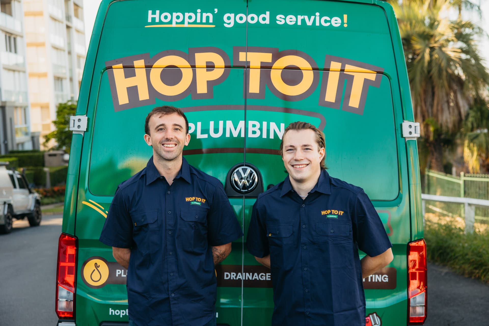 Two Hop To It roof plumbers in branded shirts standing in front of a green van